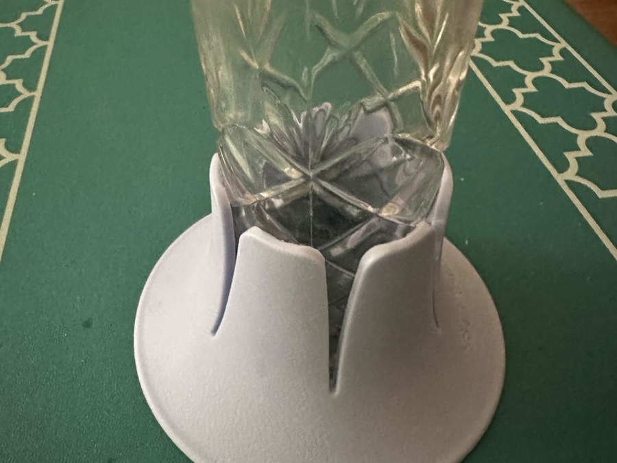 A pale blue-grey drink base holding a cut-crystal tumbler of water on a green felt table with a white trellis-pattern border