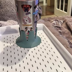 A sage-green drink base holding a stainless tumbler wrapped in a dog-print design, standing on a white slotted serving tray on a soft grey blanketed surface in a living room