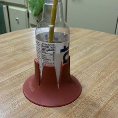 A clear glass drink bottle with a yellow straw sitting in a brick-red drink base on a light oak kitchen table, pale green cabinets and a kitchen counter behind